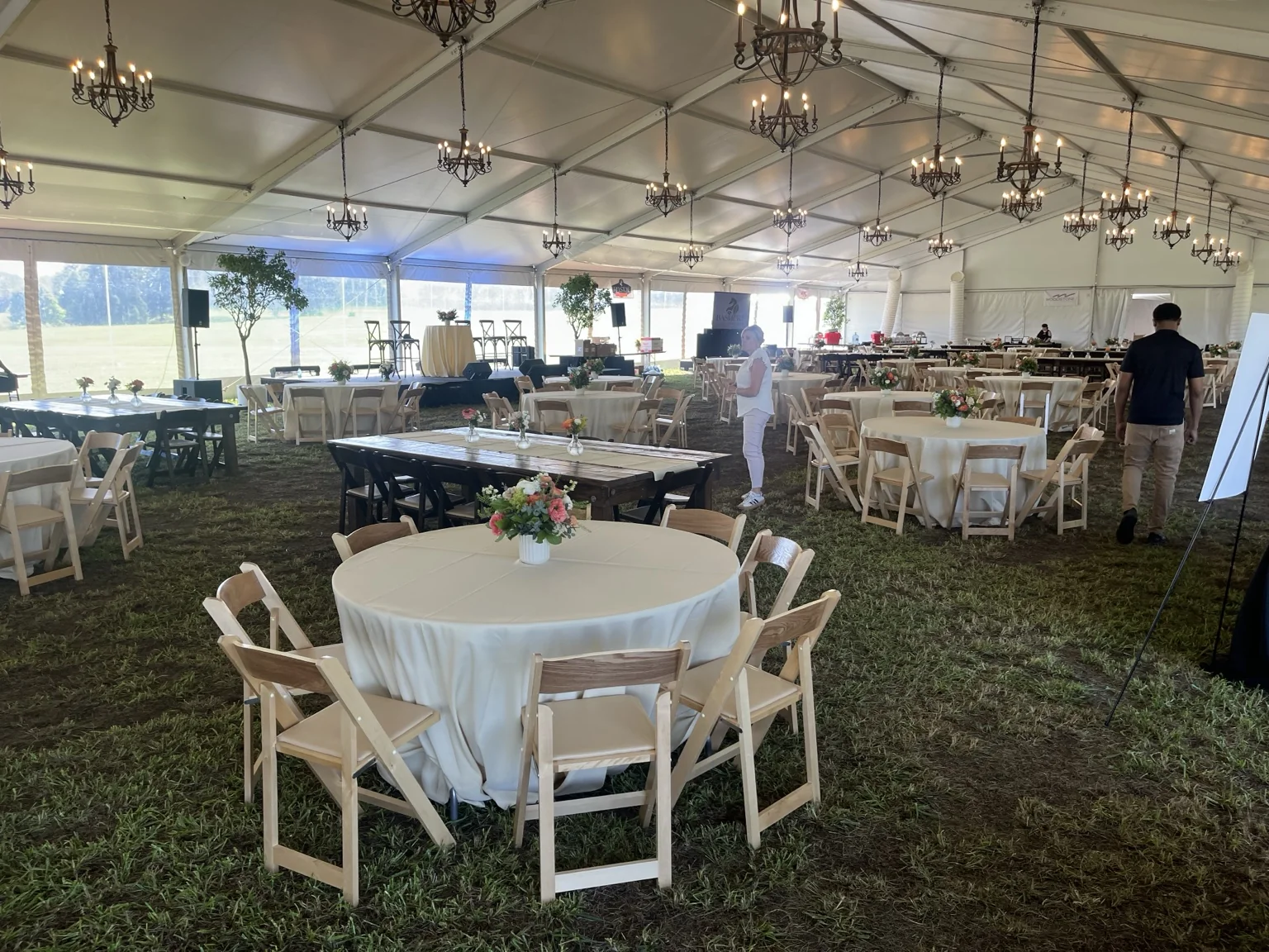 Tented wedding reception with ivory linens, natural wood chairs, floral centerpieces, chandeliers and mixed farm tables arranged across a grass floor.