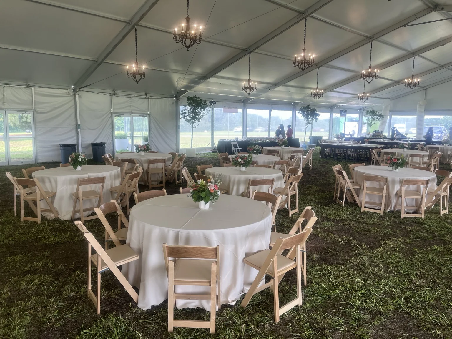 Wide tented wedding reception layout with ivory round tables, wood chairs, floral centerpieces and chandeliers overhead.
