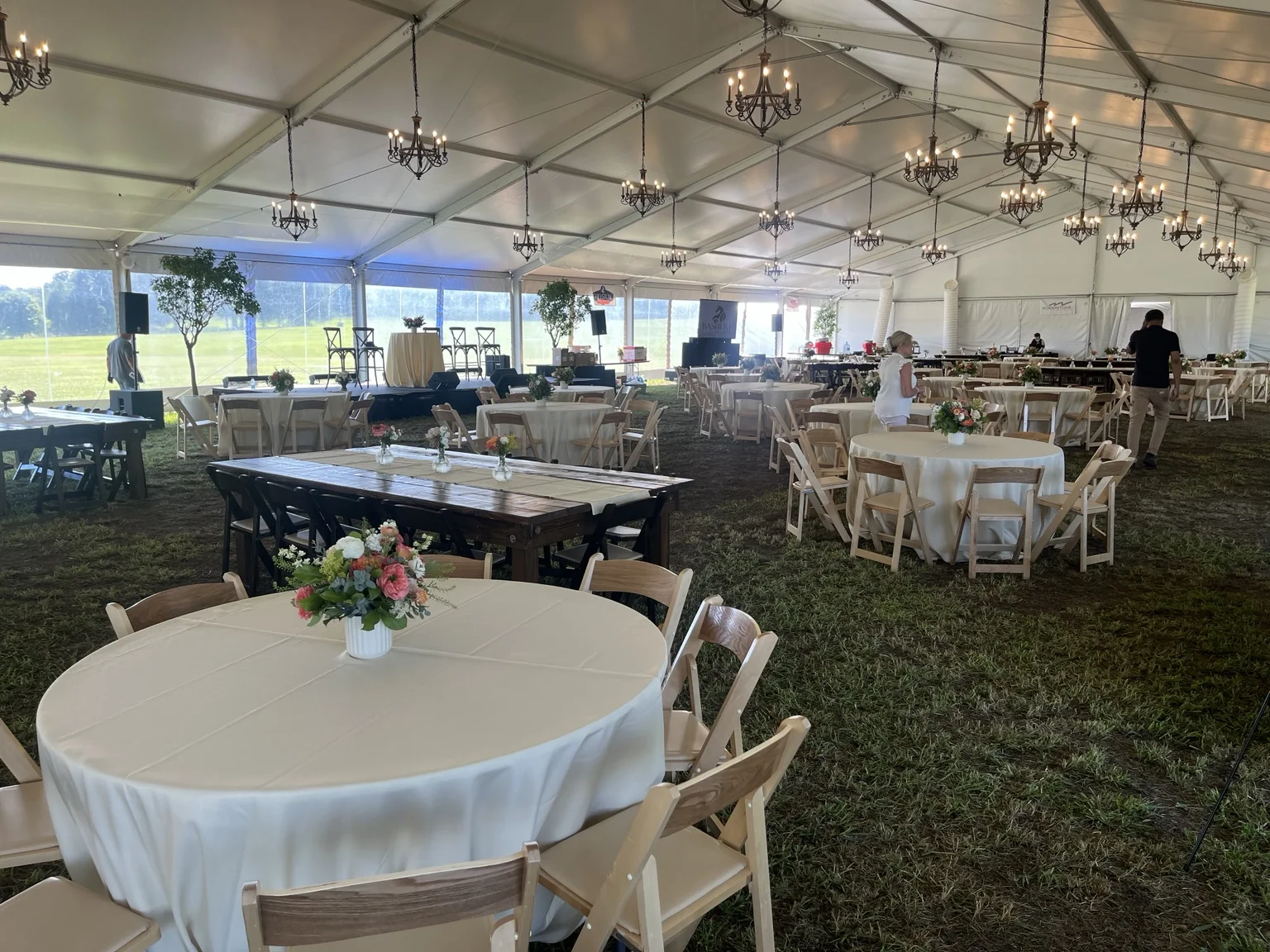 Wide view across a tented wedding reception with chandeliers, ivory tables, floral centerpieces, farm tables and open lawn visible beyond the clear tent walls.