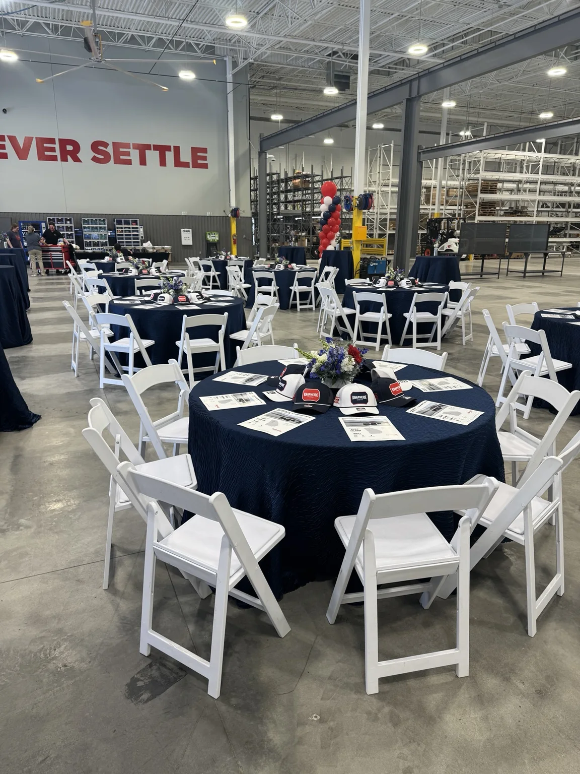 Corporate grand opening seating layout with navy linens, white chairs, branded table details and balloon columns inside an industrial venue.
