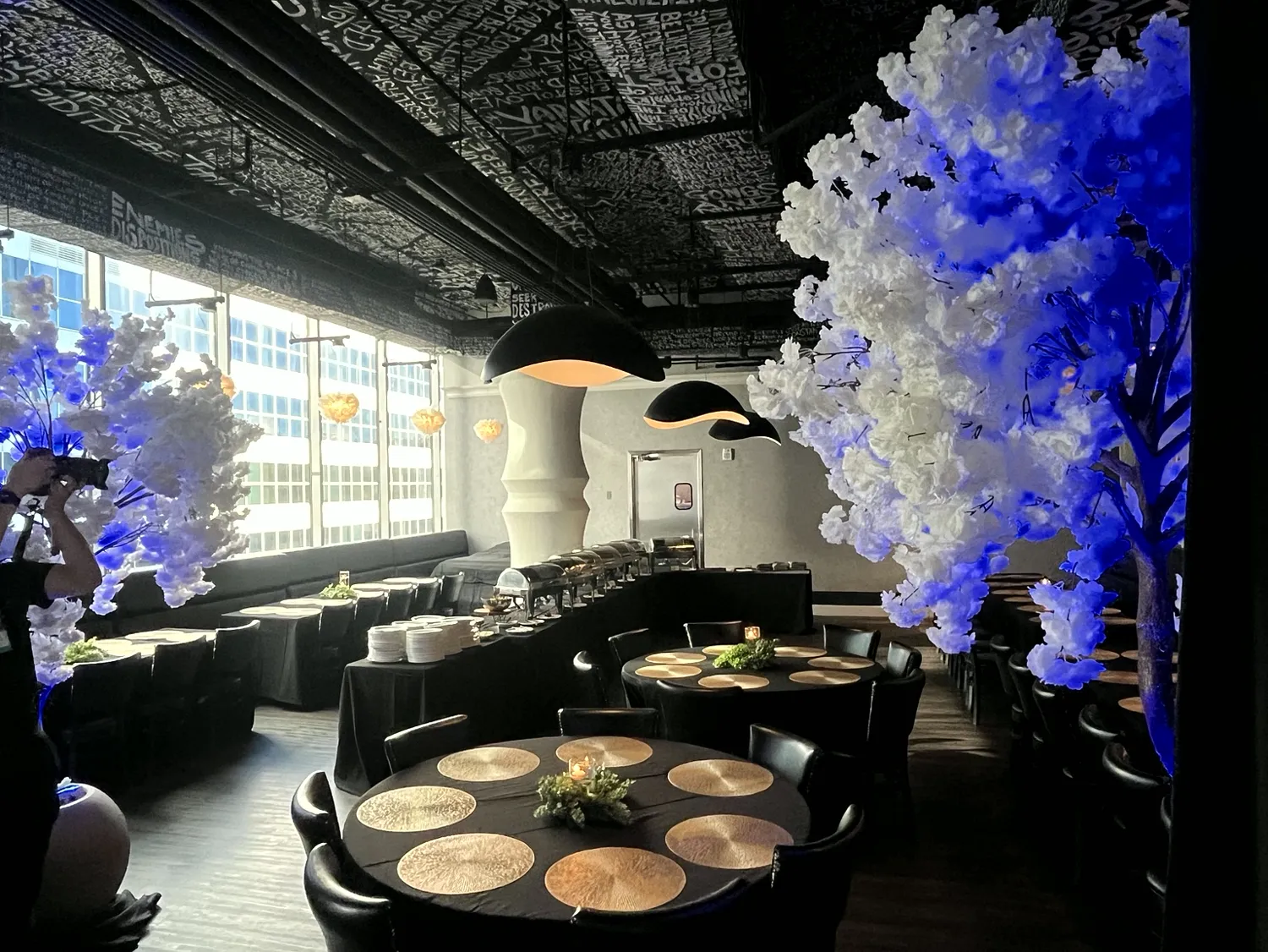 Dark reception room prepared with black linen tables, gold place settings, buffet service, sculptural lighting and illuminated white floral trees.